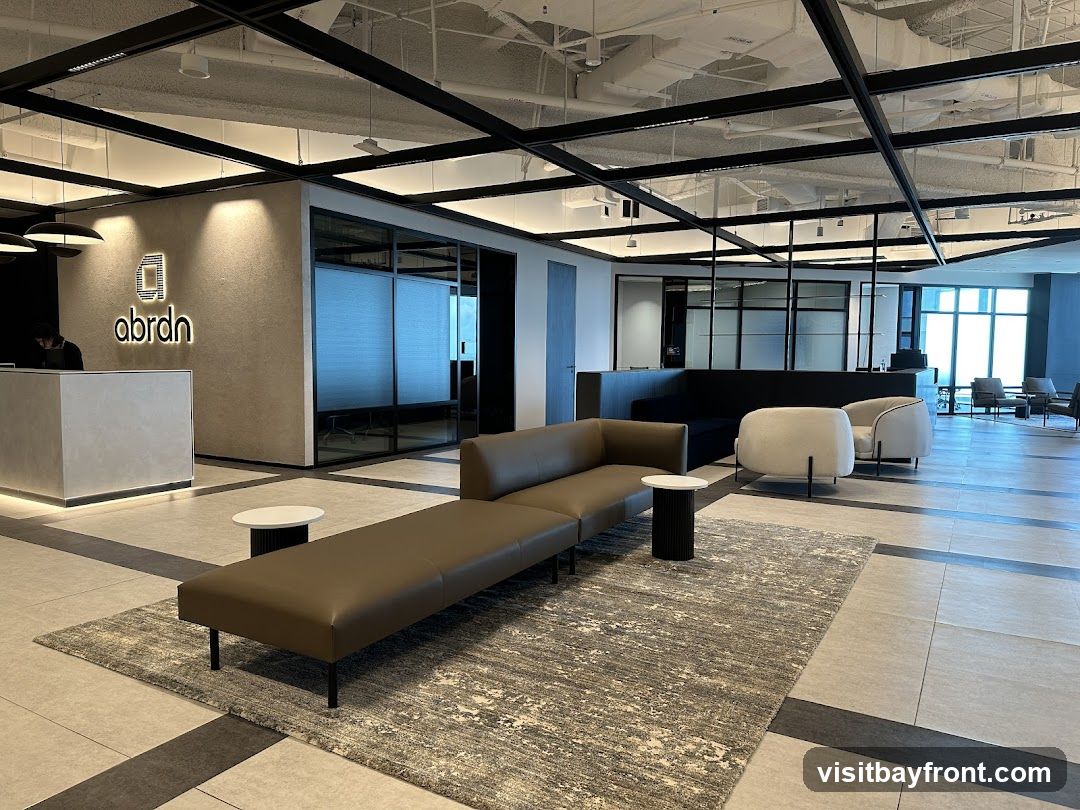 A contemporary office reception area with a sleek reception desk, comfortable seating, and a branded logo on the wall. The space features a long, modular brown leather sofa, a white accent chair, and a patterned rug. The ceiling has exposed ductwork and modern lighting fixtures. Large windows in the background offer a view of the city.
