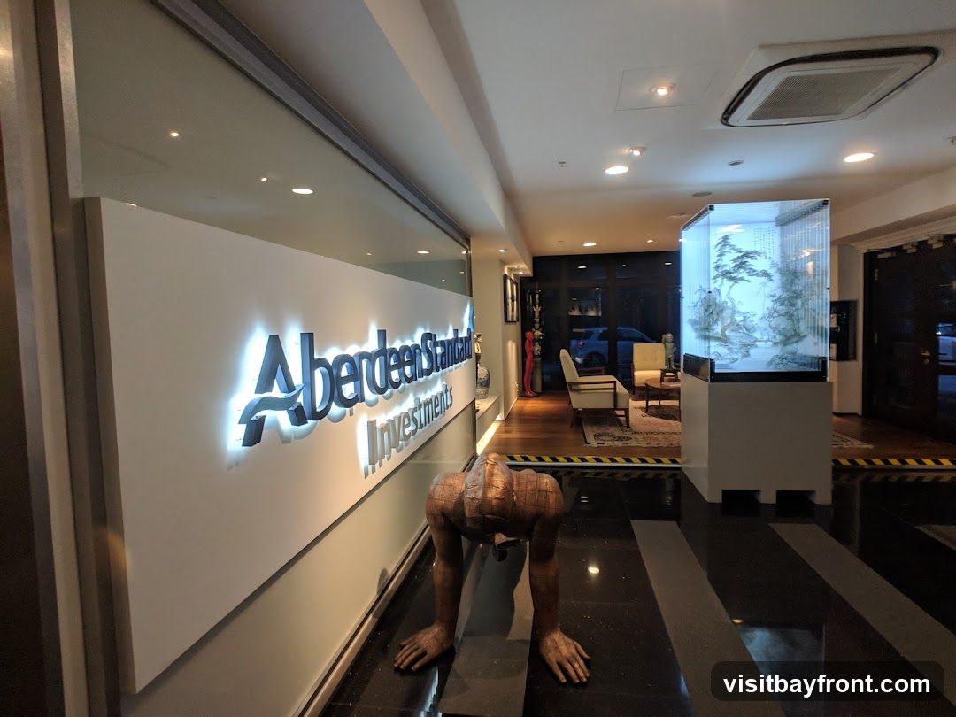 A well-lit lobby with a prominent illuminated sign for Aberdeen Standard Investments. In the foreground, a sculpture of a person in a crawling or stretching pose. To the right, a modern display case with a miniature landscape scene. Further back, seating areas with chairs and a coffee table, and a glimpse of an exterior through a doorway.