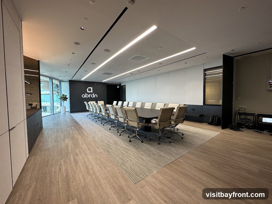 A spacious and modern conference room with a large oval table surrounded by numerous executive chairs. The room features sleek, minimalist design elements, including integrated lighting, a dark accent wall with the 'abrdn' logo, and large windows offering a view of a cityscape. The flooring transitions from wood-look laminate to carpet under the table. Equipment racks and monitors are visible in the background.