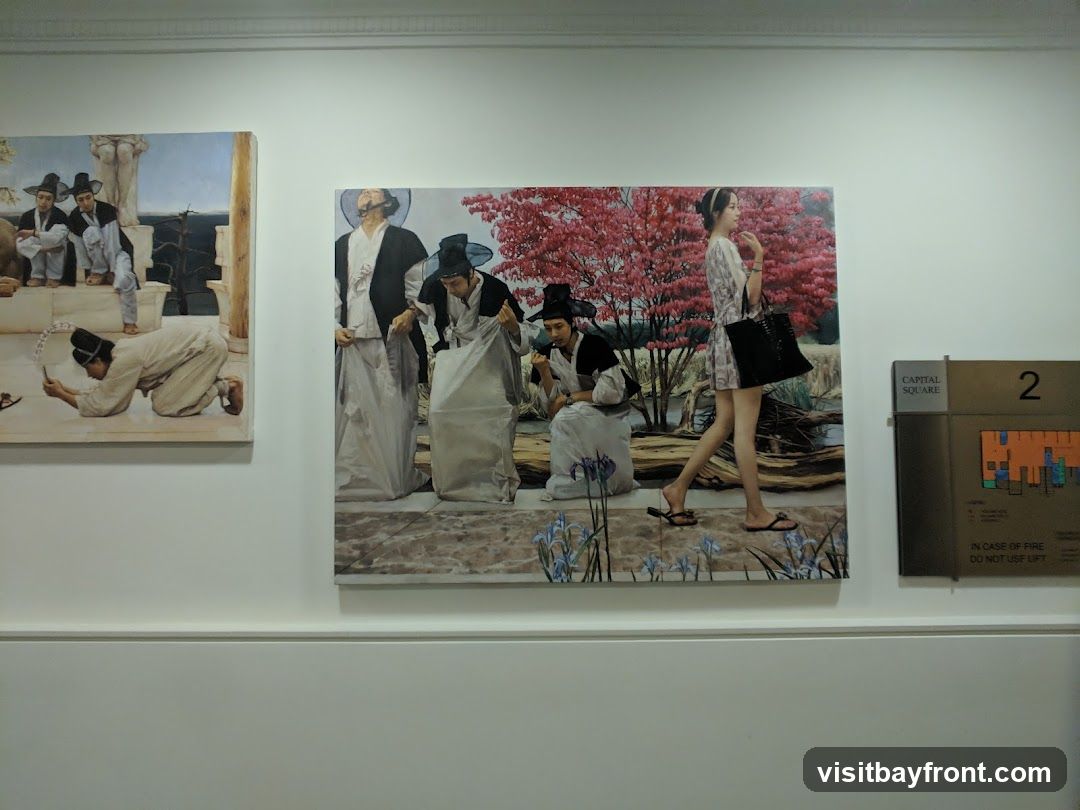 Two paintings are displayed on a white wall. The painting on the left depicts several figures in traditional attire, with one figure kneeling and holding a fan. The painting on the right shows a group of people in similar traditional clothing, with a woman in modern attire walking past them. A sign with "CAPITAL SQUARE" and the number "2" is visible to the right of the paintings.