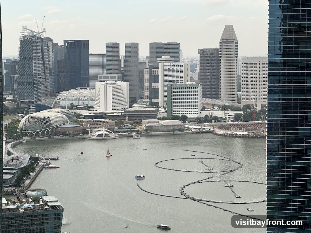 A panoramic view of a modern cityscape with a large body of water in the foreground. Several skyscrapers of varying architectural styles dominate the skyline. On the waterfront, distinctive buildings with unique designs are visible, including a domed structure resembling a durian fruit and a building with a tiered, stepped facade. The water is dotted with small boats and a sailboat, and circular floating structures are present. The overall scene suggests a bustling urban environment with a prominent waterfront.
