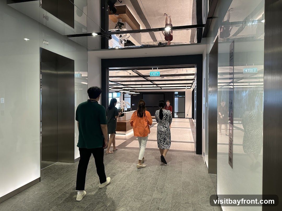 A group of people walk down a modern office hallway with elevators on the left and a reception area visible in the distance. The ceiling has a reflective surface, showing upside-down reflections of people and the interior. Green exit signs are visible on the ceiling.