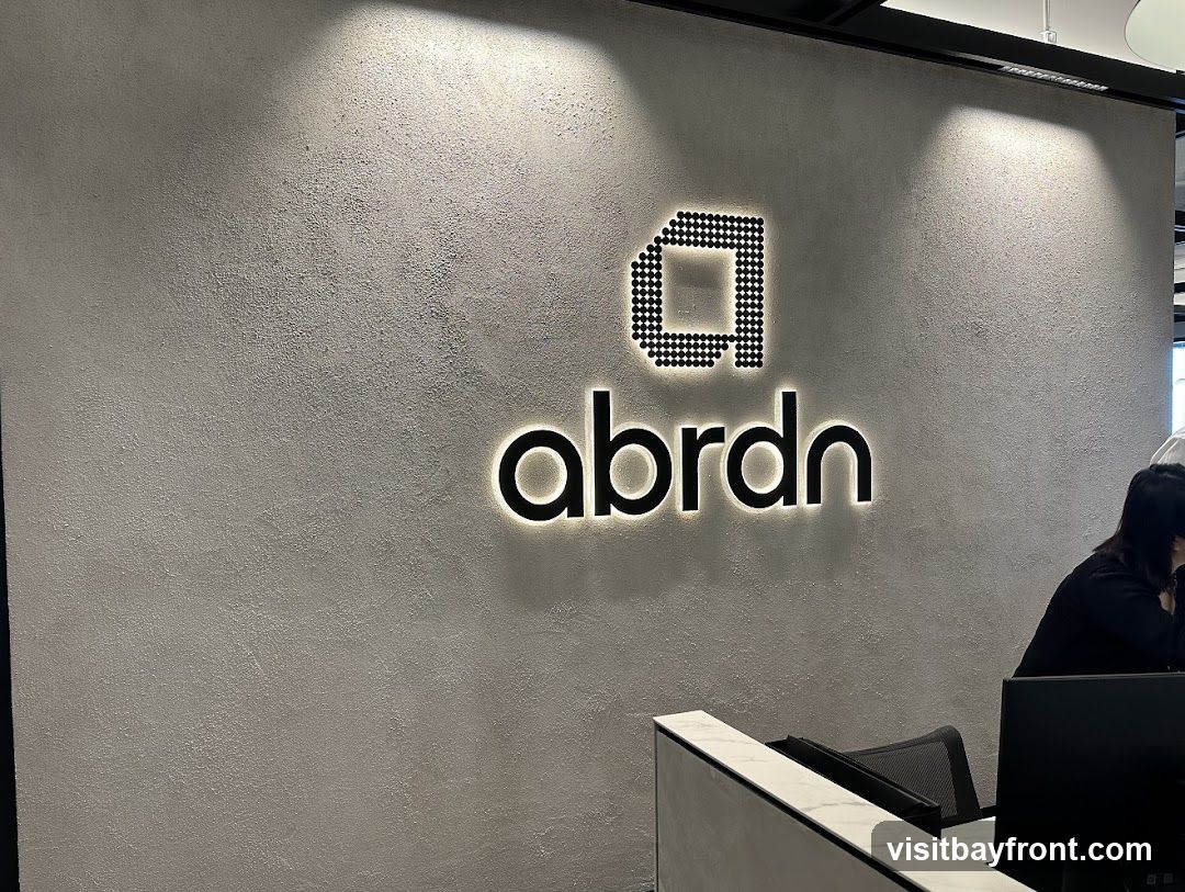 A modern office interior featuring the Abrdn logo illuminated on a textured grey wall. The logo consists of a stylized 'a' formed by dots above the word 'abrdn' in a sleek, illuminated font. To the right, a person is seated at a reception desk with a computer monitor.
