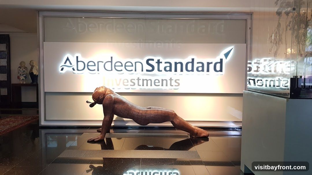 A sculpture of a person in a plank position is displayed in front of a large illuminated sign that reads "Aberdeen Standard Investments". To the left of the sign, there are two small figurines of an elderly couple. The floor is highly reflective, showing the reflections of the sign and the sculpture.