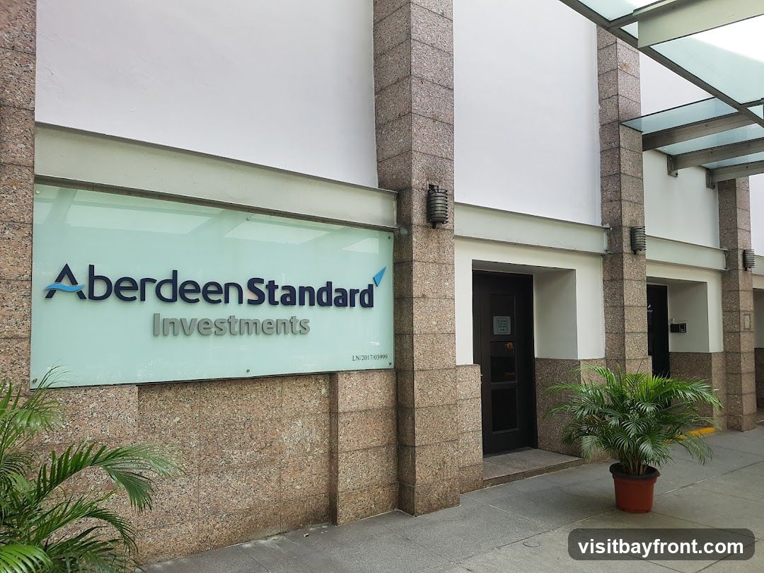 The exterior of a building with a large glass sign displaying "Aberdeen Standard Investments" in blue and silver lettering. The building has stone-clad pillars and white walls. There are two dark doors visible, one with a sign on it. Potted plants are placed outside the entrance.
