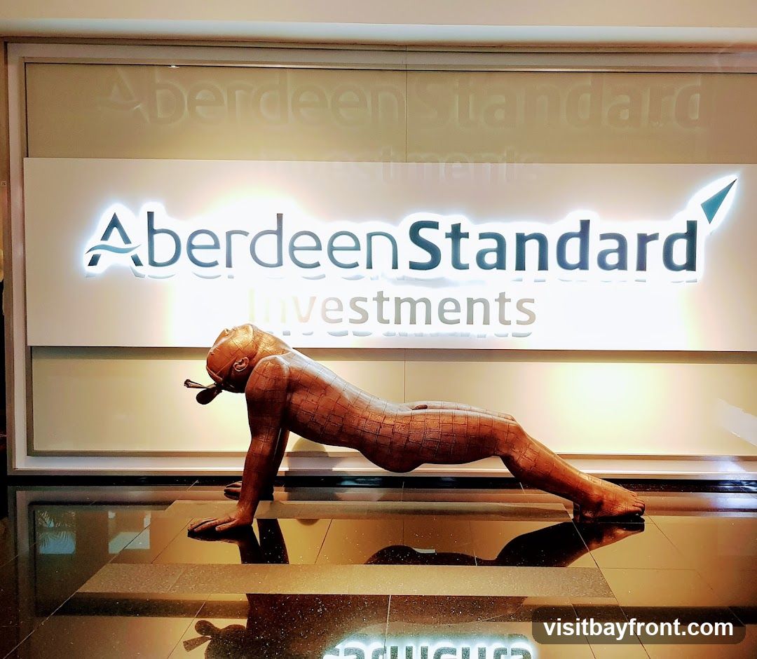 A bronze sculpture of a reclining figure is displayed in front of a backlit sign that reads "Aberdeen Standard Investments". The sign has a blue wave graphic and a teal arrow pointing upwards. The floor is highly reflective, showing a mirrored image of the sculpture and the sign.