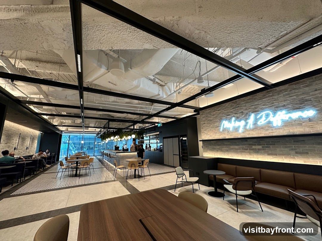 The image shows the interior of a modern cafe with a long bar, seating areas, and a neon sign on the wall. The ceiling is exposed with ductwork and lighting. The floor has a tiled pattern. There are tables and chairs arranged for dining.