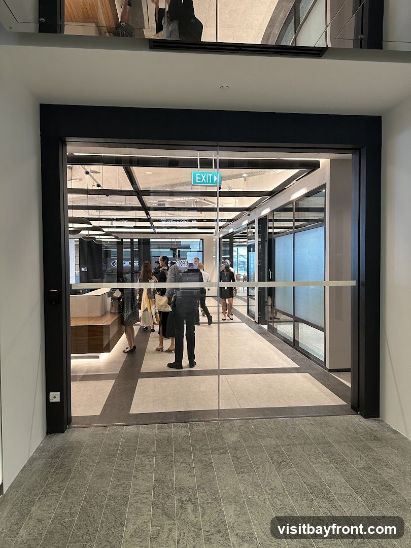 A view through glass doors into a modern office space. People are seen walking down a hallway with a reception desk on the left. The ceiling features a grid of lights and panels. An "EXIT" sign is visible above the doorway.
