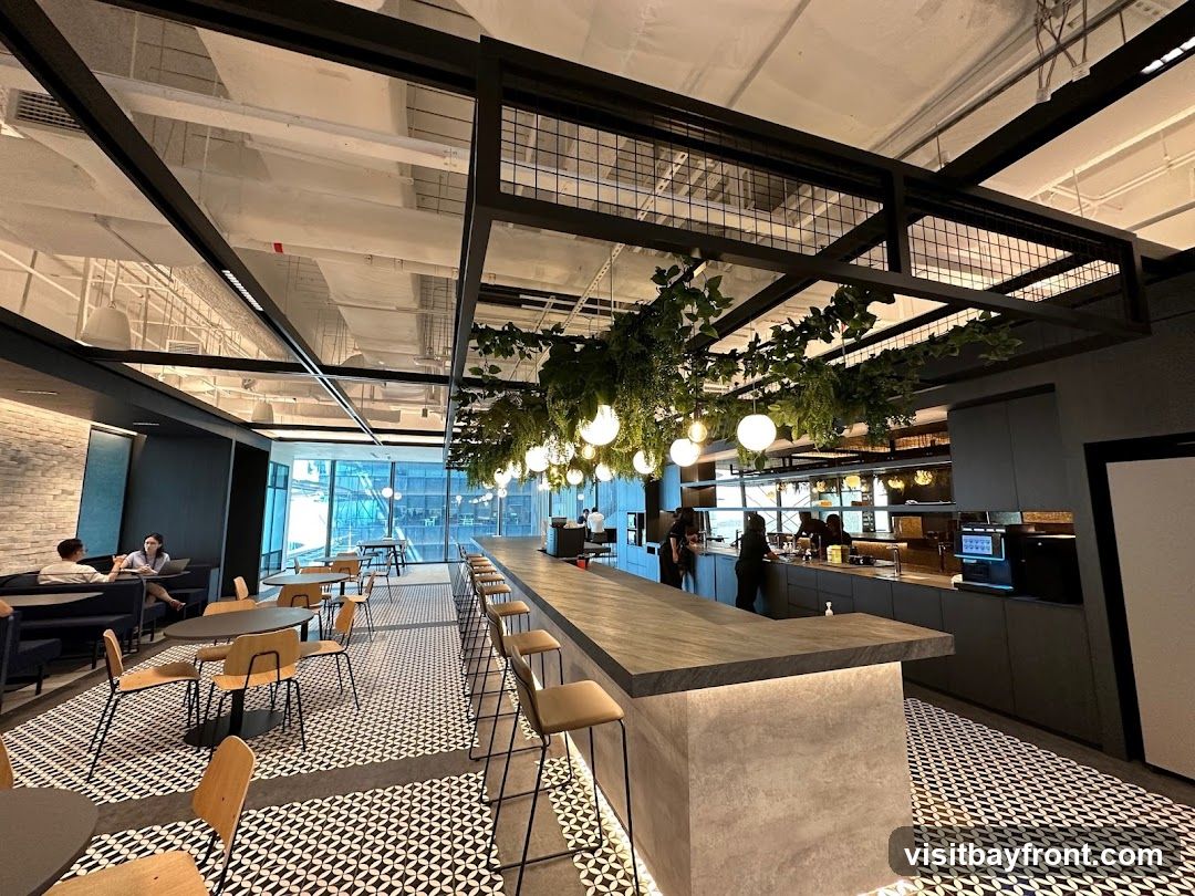 An interior view of a modern office break room with a long counter bar, seating areas, and a kitchen area. The space features a black and white patterned floor, hanging plants, and pendant lights. Several people are visible in the background, some seated at tables and others working behind the counter.