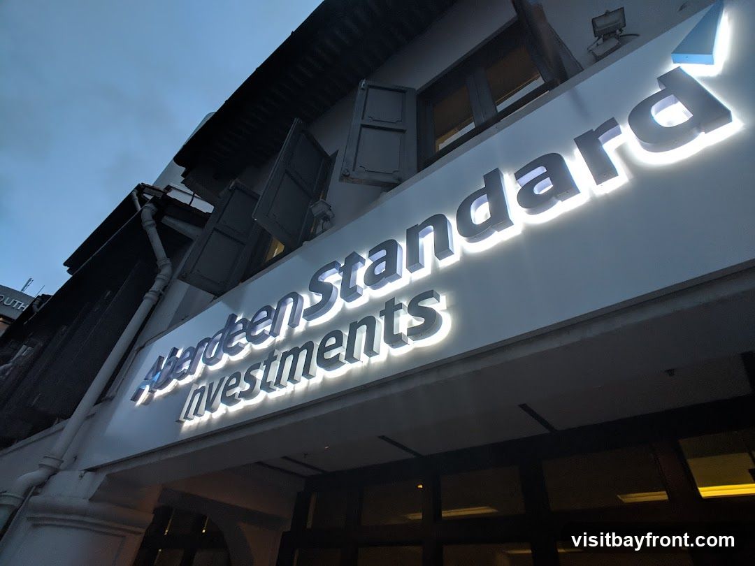A close-up, low-angle shot of a lit sign for "Aberdeen Standard Investments" mounted on the exterior of a building. The sign is white with illuminated lettering. Above the sign, traditional shutters are open on windows. The sky is dark blue, suggesting dusk or night.