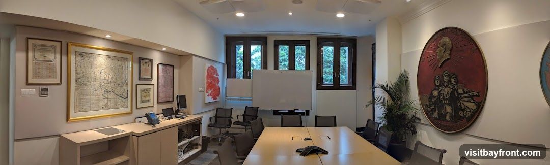A panoramic view of a conference room featuring a large table with chairs, whiteboards, and framed maps and artwork on the walls. There is also a desk with office equipment and a plant in a pot.