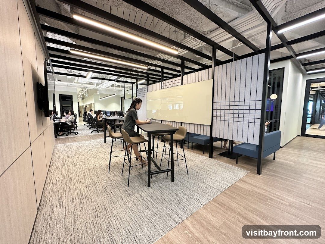 A contemporary office interior featuring a high-top table with chairs in the foreground, where a woman is seated and looking at her phone. Behind her, a large whiteboard is mounted on a partition. Further back, rows of desks with people working are visible, along with lounge seating areas. The space is illuminated by modern linear lighting and has a mix of carpeted and wood-look flooring.