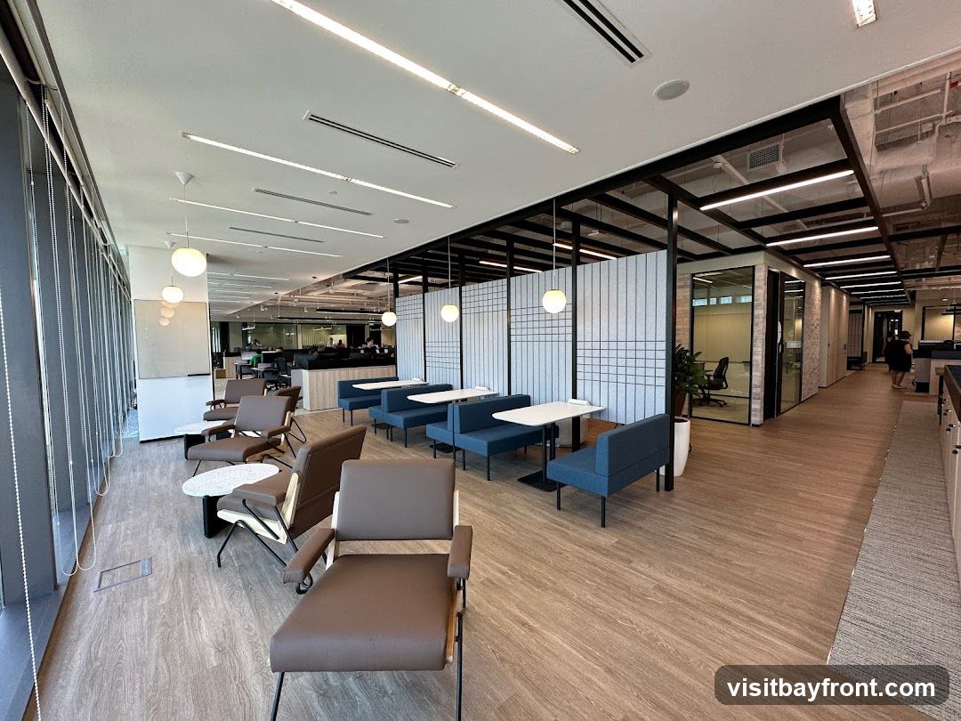A contemporary office break area with comfortable seating, including plush armchairs and booth-style seating with tables. The space features large windows with blinds, modern lighting fixtures, and a clean, minimalist aesthetic. Partitions with a textured pattern divide sections of the room, and glass-walled meeting rooms are visible in the background. The flooring is a light wood laminate.