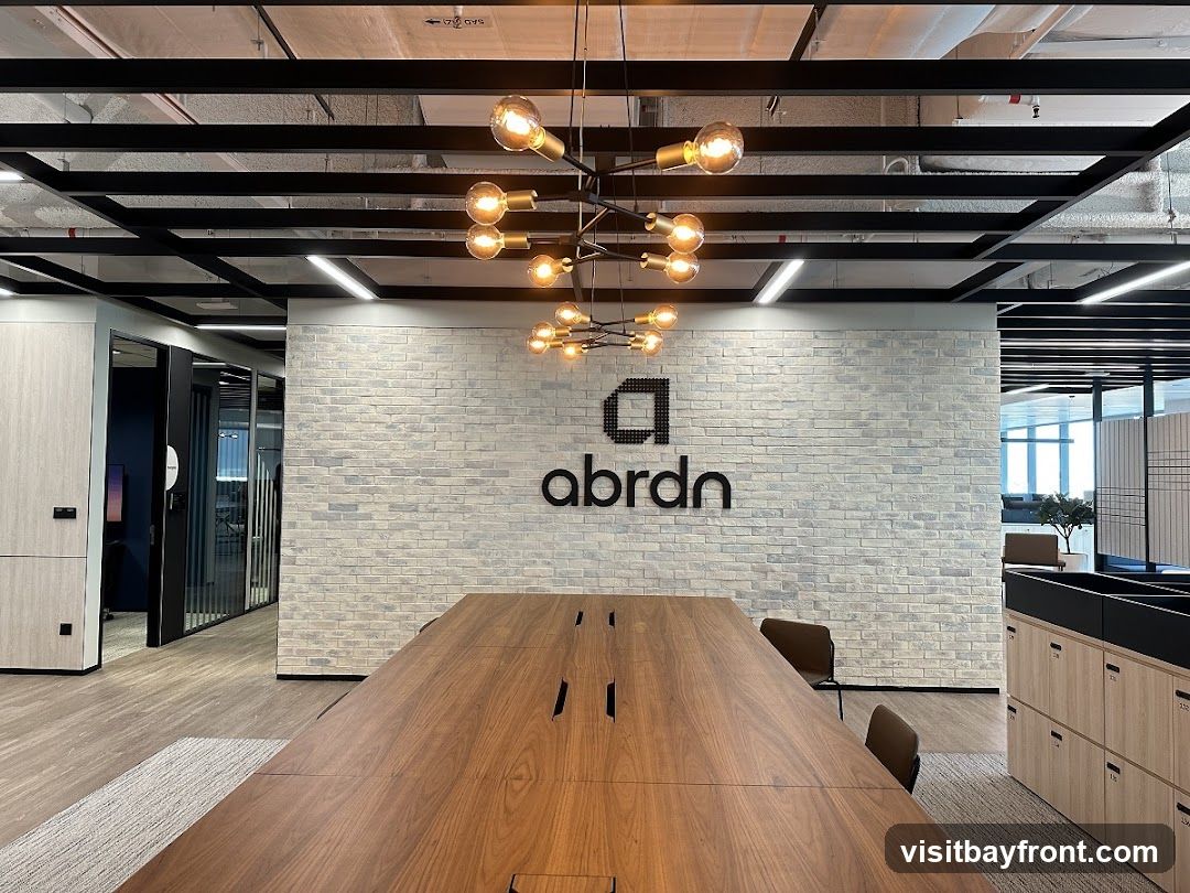 A modern office interior featuring a long wooden conference table in the foreground. Behind the table, a white brick wall displays the black 'abrdn' logo. The ceiling has exposed black beams and industrial-style lighting fixtures. To the left, glass-partitioned offices are visible, and to the right, there are wooden cabinets with numbered drawers. The overall ambiance is sleek and professional.