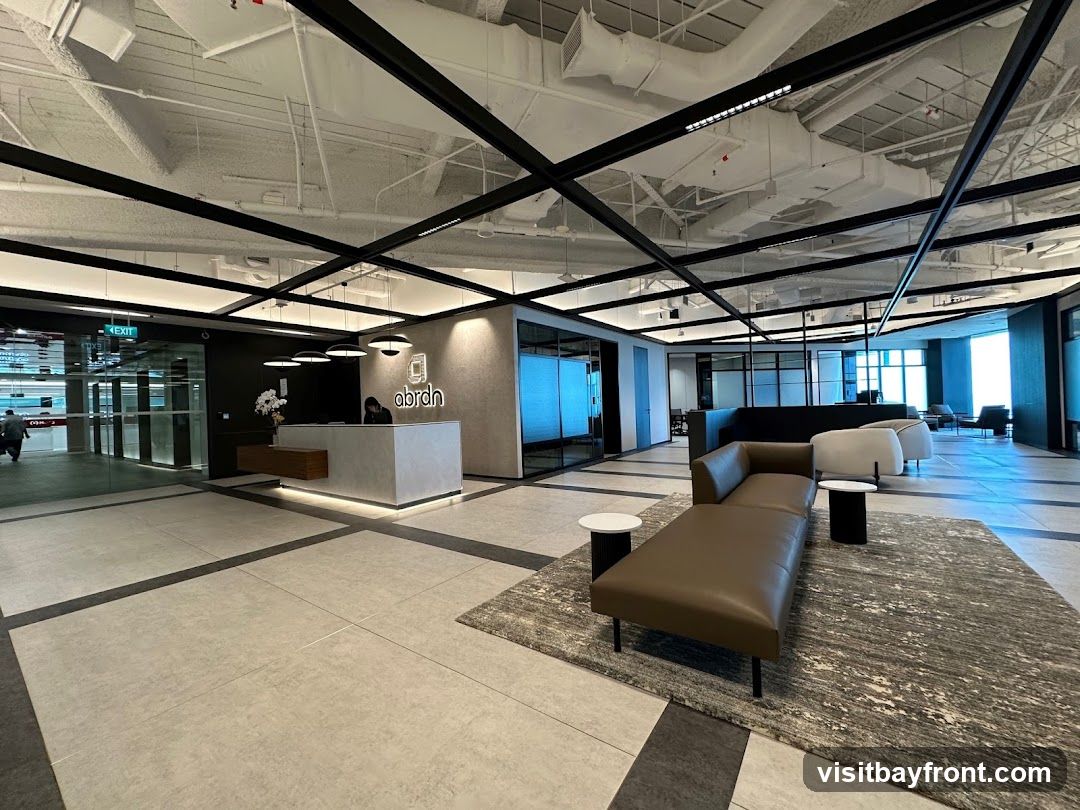 A modern office reception area with a grey reception desk featuring the 'abrdn' logo. To the right, a brown leather chaise lounge sits on a patterned rug with small side tables. The space has an open-plan design with glass-partitioned offices and a view of the city through large windows.