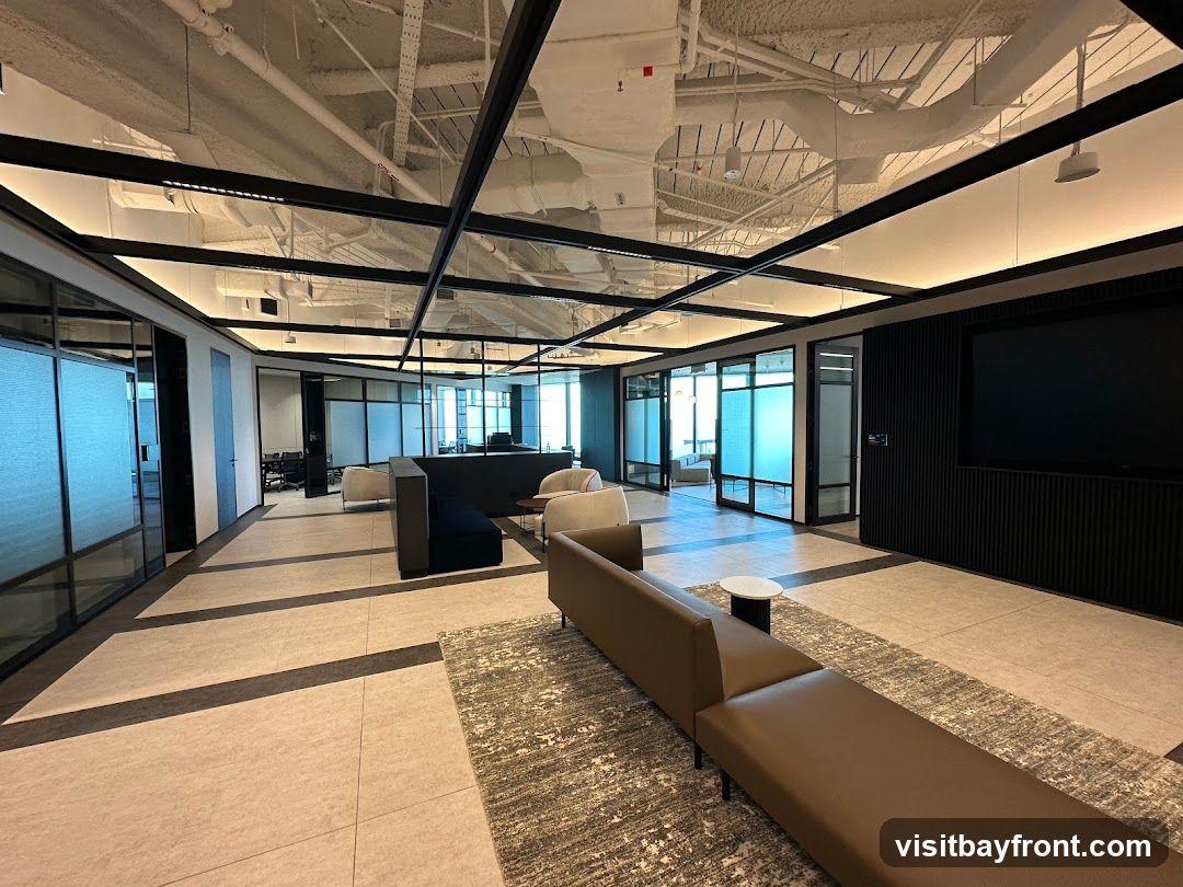 A spacious and modern office lounge area with contemporary furniture. The space features a long, L-shaped brown leather sofa, a dark blue sofa, and several armchairs. The flooring is a light-colored tile with dark accent strips, complemented by a patterned area rug. Glass-walled offices and meeting rooms are visible in the background, with frosted glass panels providing privacy. The ceiling has exposed industrial elements and a grid of modern lighting fixtures. A large black screen is mounted on a textured black wall on the right side of the room.