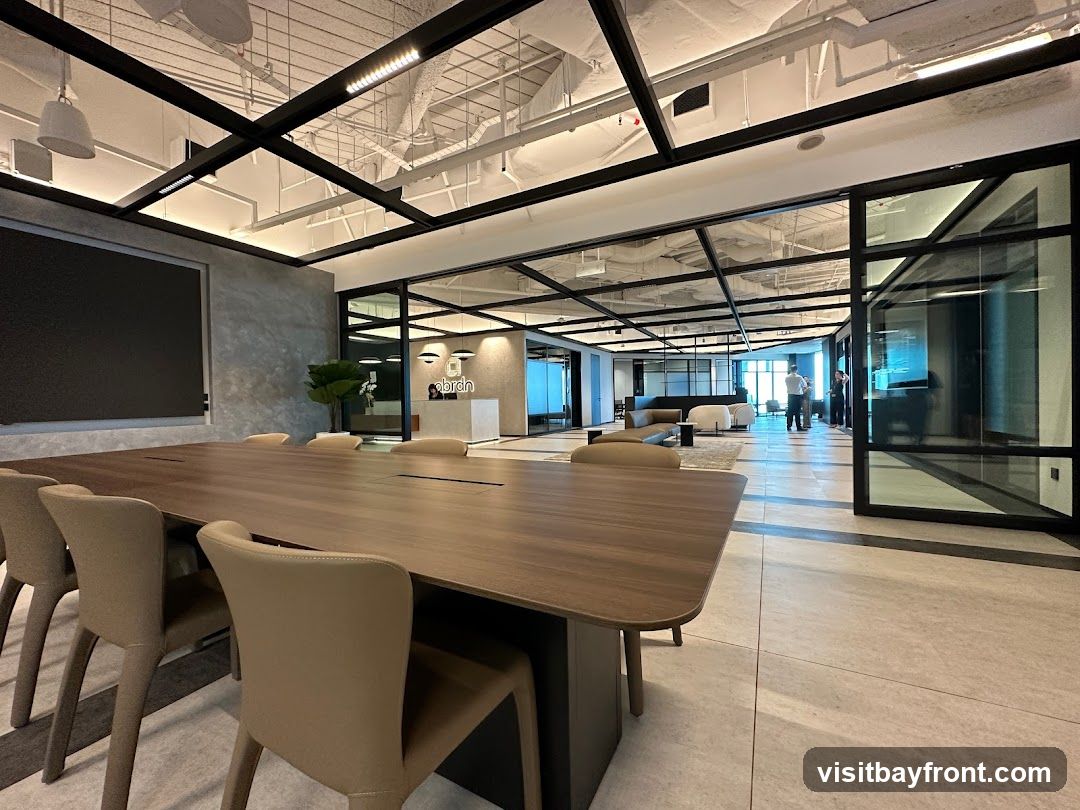 A spacious and modern office meeting room with a large wooden conference table and chairs in the foreground. In the background, through glass partitions, a reception area with a logo and a lounge area with contemporary furniture are visible. The ceiling features exposed ductwork and lighting.