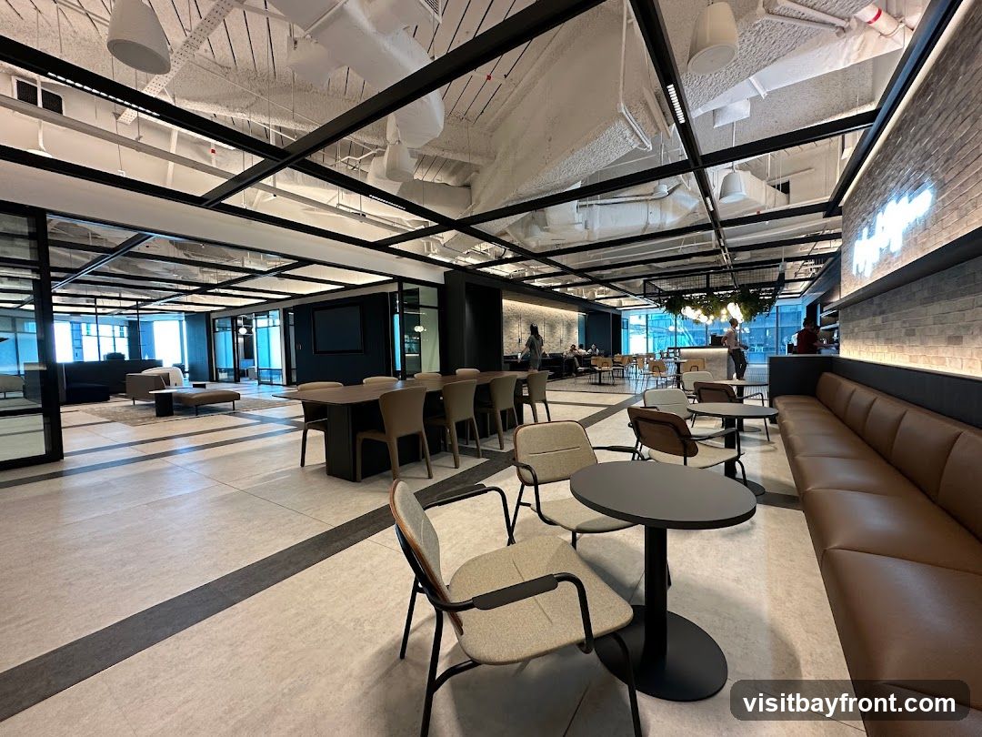 A spacious and modern office break room with various seating areas, including a long leather banquette, small round tables with chairs, and a large conference table. The space features exposed ceilings with black beams and ductwork, industrial-style lighting, and a brick accent wall with a neon sign. Large windows offer views of the city, and plants add a touch of greenery.