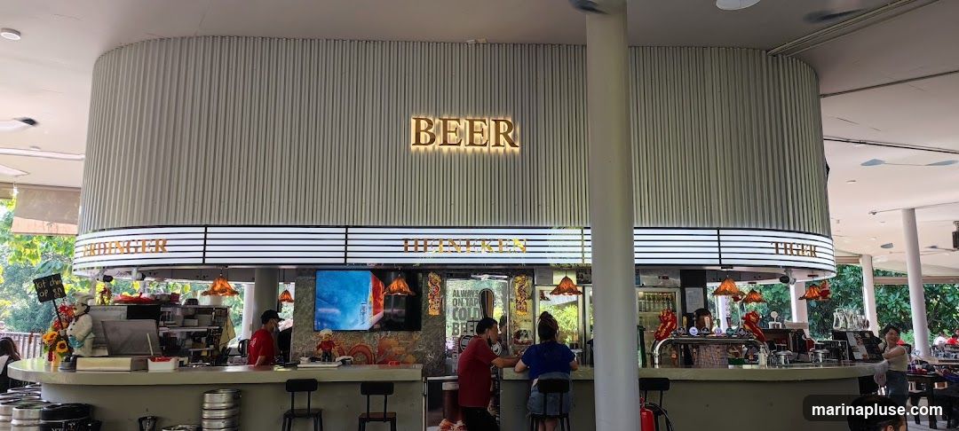 A long bar with a prominent illuminated sign that reads "BEER" above it. Below the beer sign, there are illuminated signs for "HEINEKEN" and "TIGER". People are seated at the bar and standing, with staff serving. There are beer taps and bottles visible behind the bar. The overall ambiance appears to be a casual outdoor or semi-outdoor bar setting.