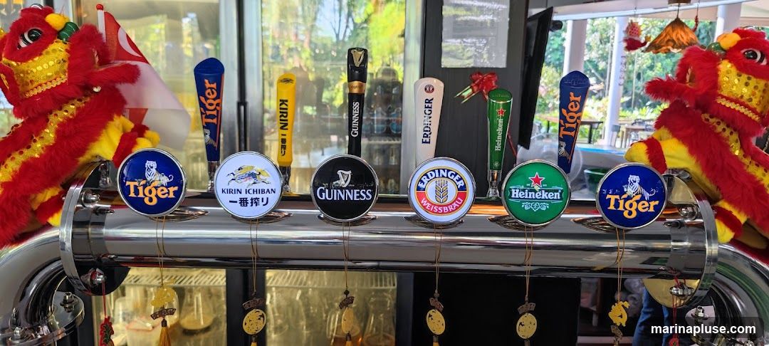 A row of beer taps with various brand logos including Tiger, Kirin Ichiban, Guinness, Erdinger, and Heineken. Decorative lion dance figures and a Singapore flag are visible on either side.