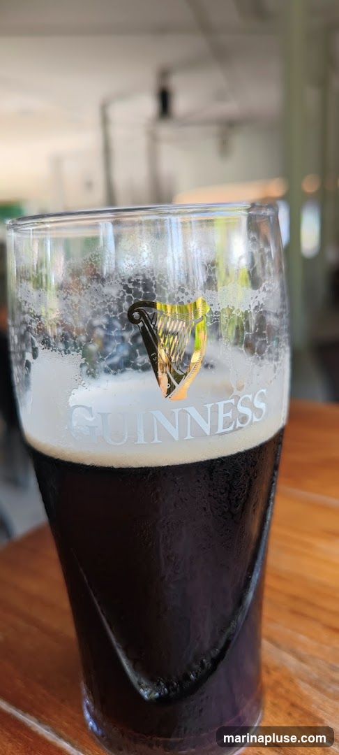 A close-up shot of a glass of Guinness beer on a wooden table. The glass has the Guinness harp logo and name printed on it. The beer has a dark color and a foamy head. The background is blurred and appears to be an indoor or outdoor seating area.