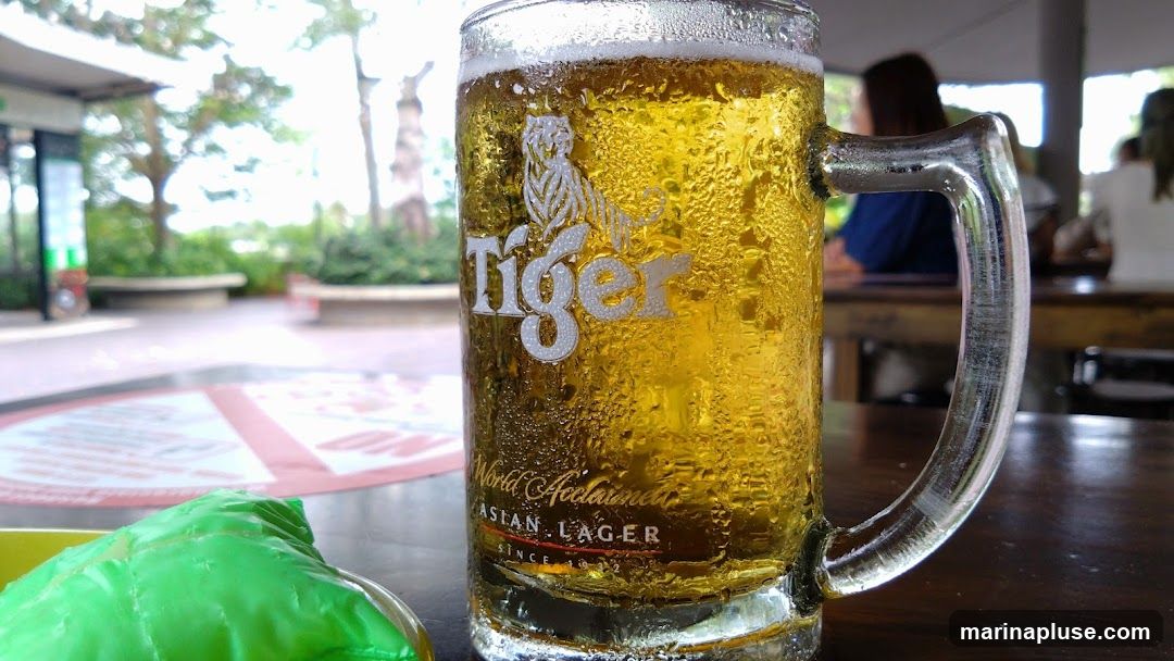 A close-up shot of a frosty mug of Tiger Asian Lager beer sits on a wooden table. The iconic tiger logo and text are visible on the glass. In the blurred background, people are seated at tables, suggesting an outdoor seating area of a restaurant or bar. A green item, possibly food, is in the foreground.