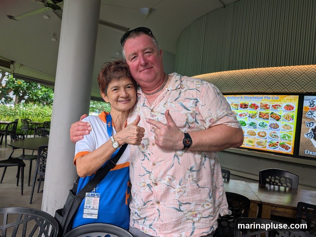 A man and a woman are posing for a photo, with the man's arm around the woman. They are both smiling and giving thumbs up. Behind them is a large digital display showing various food dishes, likely from a restaurant. The display has text that reads "Seafood & Steamboat Pte Ltd" and appears to be a menu or advertisement. Tables and chairs are visible in the background, suggesting an outdoor dining area.