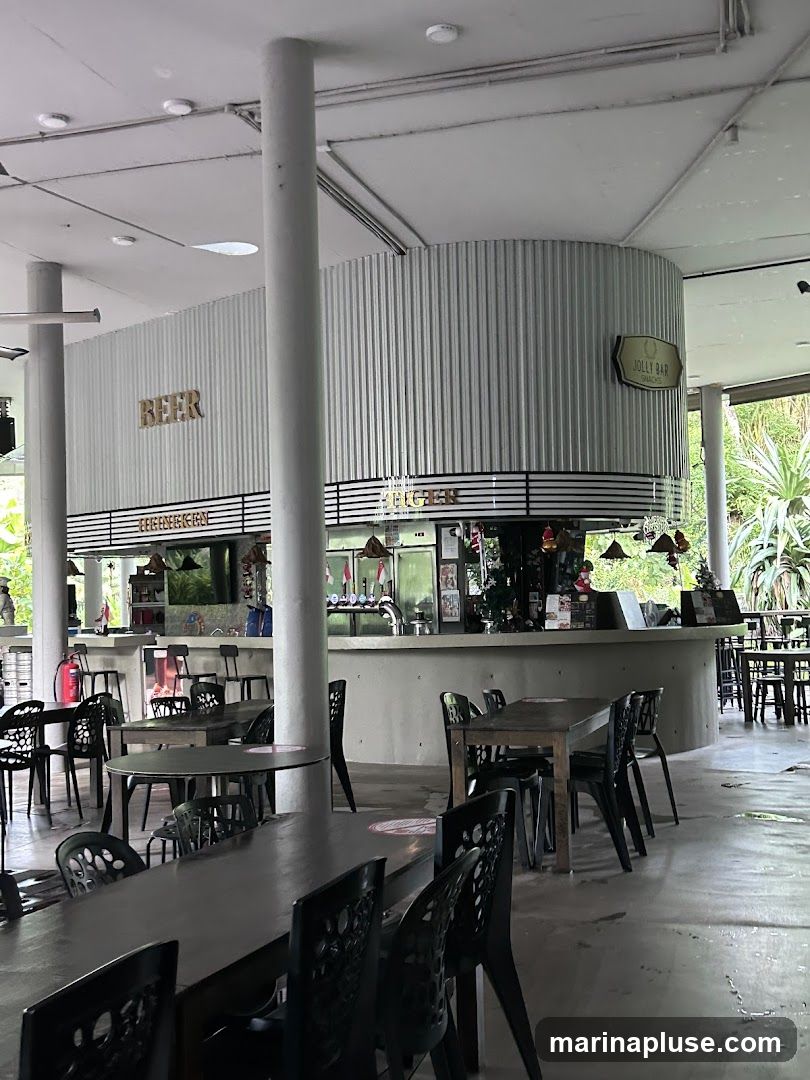 The image shows the interior of a bar with a curved counter and seating areas. The counter has beer taps and a sign that says "JOLLY BAR SNACKS". There are tables and chairs set up in front of the counter and in the foreground. The overall ambiance is casual and open-air, with greenery visible in the background.