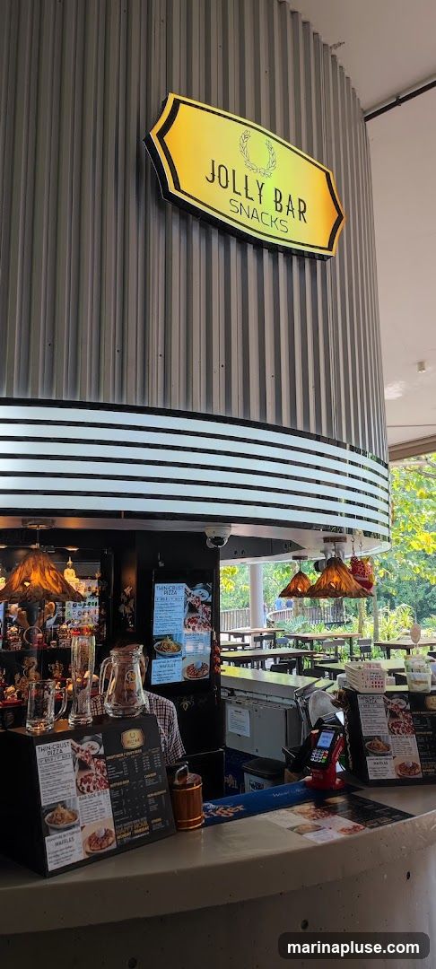 A counter area with a sign that reads "JOLLY BAR SNACKS". There are menus displayed, including one for "THIN-CRUST PIZZA" and "WAFFLES". Various drinks and snacks are visible behind the counter. The area appears to be an outdoor or semi-outdoor seating area with tables and chairs visible in the background.