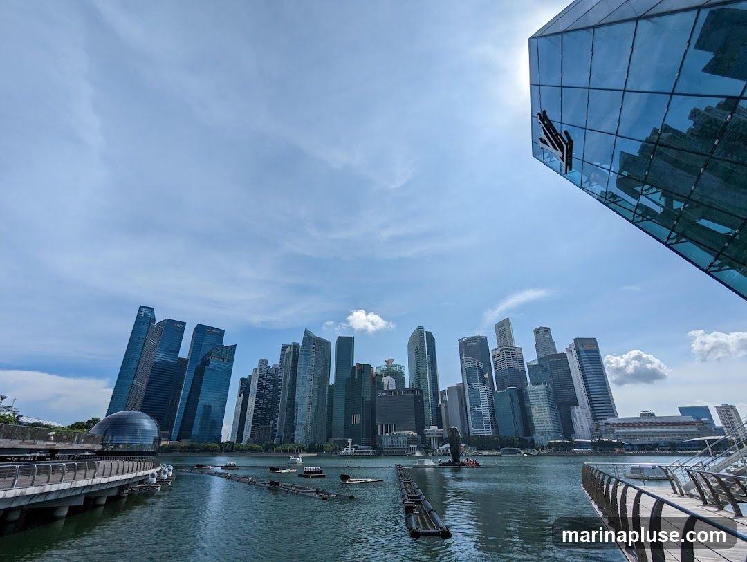 A wide-angle view of a modern city skyline with numerous skyscrapers of varying heights and architectural styles, reflected in the calm water below. A walkway with railings is visible in the foreground on the right, and another on the left. The sky is blue with scattered white clouds, and the sun is bright, casting a glare on a glass-covered building in the upper right corner. Several boats and floating structures are present on the water.