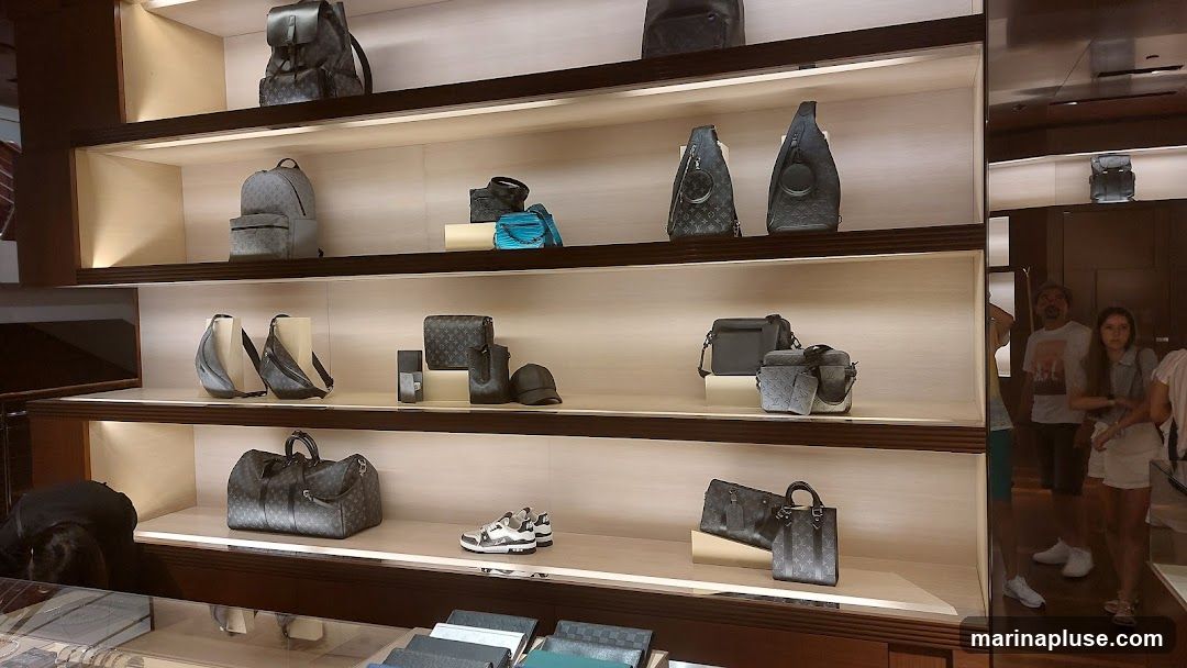 A well-lit display shelf in a luxury store showcases various handbags, backpacks, wallets, and a pair of sneakers. The items are arranged on light-colored shelves with dark wood trim. In the background, people are seen browsing the store.