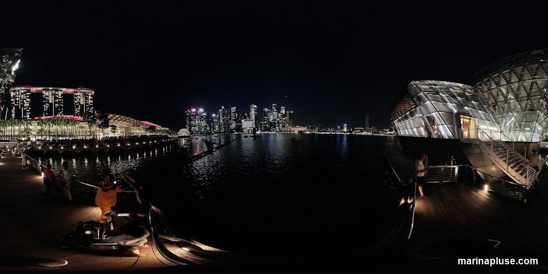 A panoramic night view of the Singapore skyline, featuring the Marina Bay Sands hotel and a modern glass building on the waterfront. People are visible on a wooden boardwalk in the foreground, some taking photos. The water reflects the city lights.