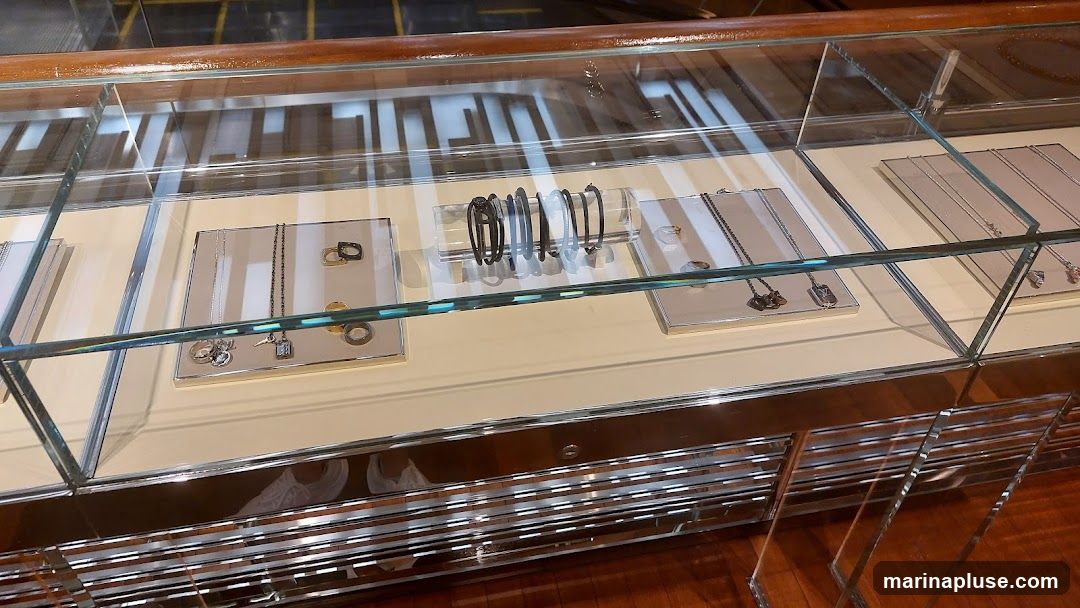 A glass display case filled with various necklaces and bracelets. The jewelry is arranged on light-colored stands within the case. In the background, an escalator is visible.