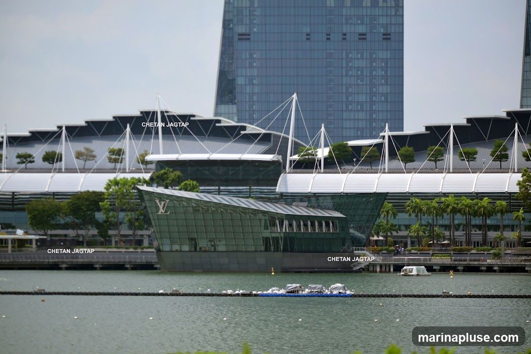 A modern, glass-fronted building with the Louis Vuitton logo visible on its facade, situated by a body of water. In the background, a large, contemporary skyscraper and a stadium-like structure with a distinctive roof design are visible.