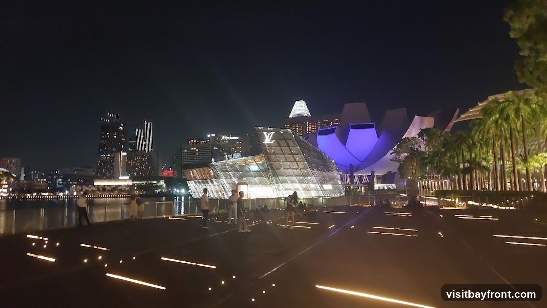 A modern, glass-faceted building with the Louis Vuitton logo prominently displayed on its facade. The building is illuminated at night, with people visible on the walkway in front of it. In the background, a cityscape with illuminated skyscrapers and a distinctive lotus-shaped building can be seen. Palm trees line the right side of the scene.