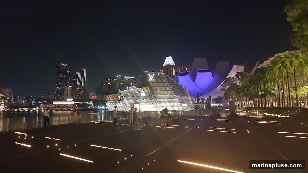 A modern, glass-faceted building with the Louis Vuitton logo prominently displayed on its facade. The building is illuminated at night, with people visible on the walkway in front of it. In the background, a cityscape with illuminated skyscrapers and a distinctive lotus-shaped building can be seen. Palm trees line the right side of the scene.