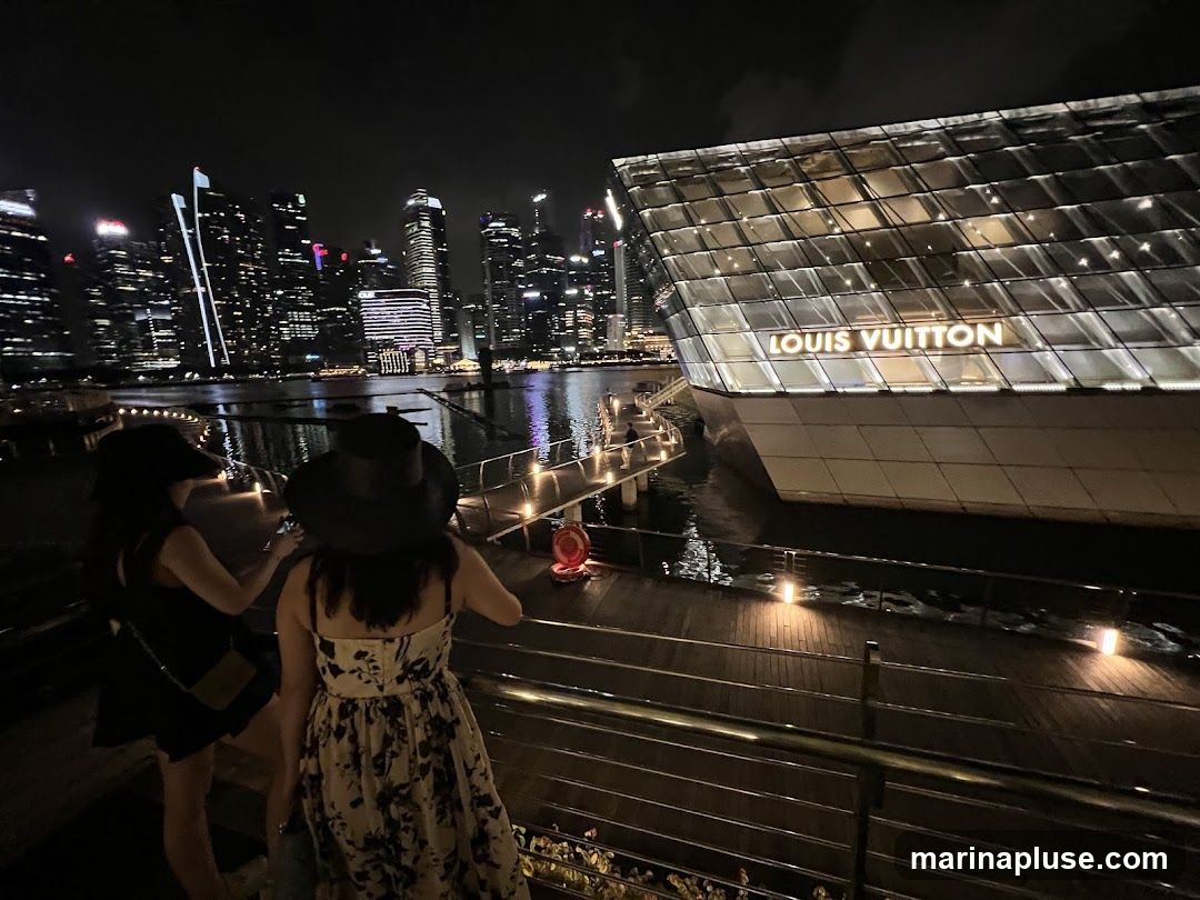 Two women stand on a wooden walkway at night, looking out at a city skyline and a modern glass building with the Louis Vuitton logo illuminated on its facade. The building appears to be situated on the water.