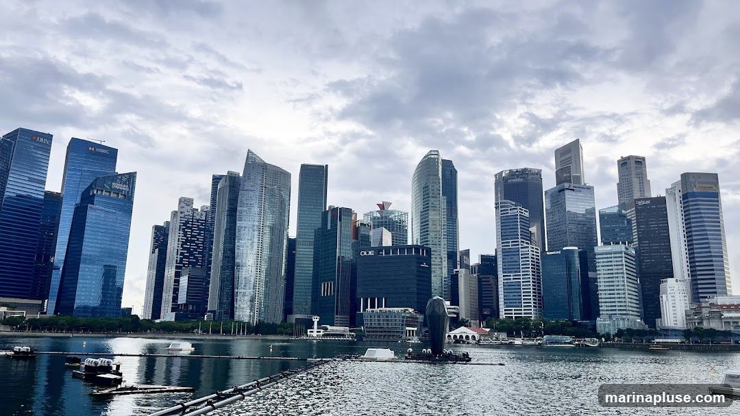A panoramic view of the Singapore skyline with numerous skyscrapers of varying heights and architectural styles. The buildings are situated along the waterfront, with a body of water in the foreground reflecting the cloudy sky and the structures. Several boats and floating platforms are visible on the water. The sky is overcast with dramatic clouds.