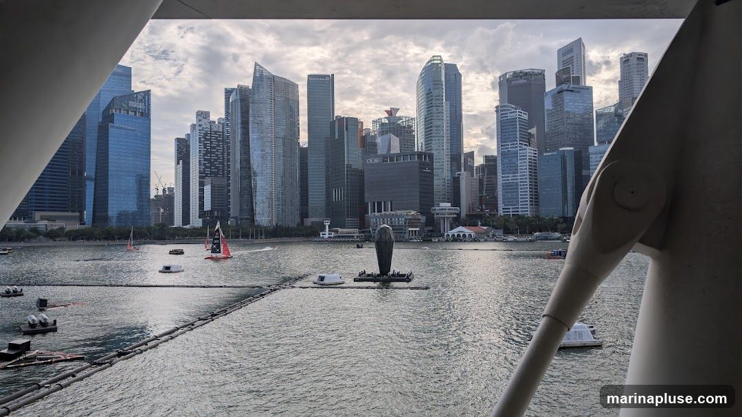A panoramic view of the Singapore skyline and Marina Bay, seen through a structural element. The image features numerous skyscrapers of varying heights and architectural styles, including buildings with glass facades. In the foreground, the water of Marina Bay is visible, with several sailboats and small floating structures. A prominent sculpture, resembling a stylized fish or rocket, stands in the bay. The sky is overcast with clouds.