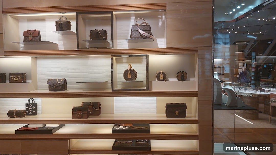 A well-lit display of various luxury handbags and accessories on shelves within a store. The items are arranged in a clean and organized manner, showcasing different styles and sizes of bags, some with the iconic monogram pattern. The reflection in the glass on the right shows a glimpse of the store's interior, including display cases and possibly mannequins or staff.