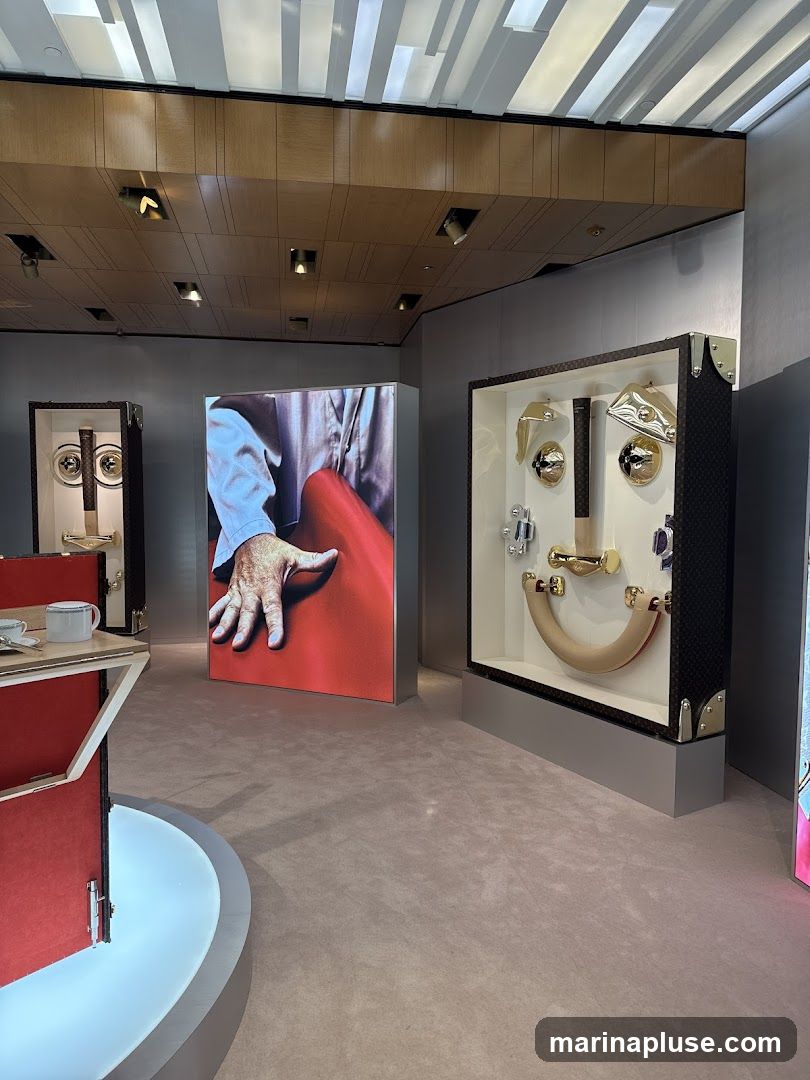 A retail display featuring artistic arrangements of hardware and imagery. In the foreground, a red display case with a white rim holds a white cup and saucer. To the left, a large illuminated panel shows a close-up of a hand reaching towards a red surface. To the right, a display case showcases various metallic hardware pieces arranged to resemble a face, set against a dark patterned background. The ceiling features a modern design with wooden panels and recessed lighting. The floor is carpeted in a neutral tone.