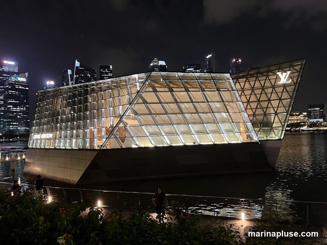 A modern, glass-encased building with a distinctive geometric design, illuminated at night. The Louis Vuitton logo is prominently displayed on one of the angled facades. The building appears to be situated on the water, with a cityscape visible in the background.