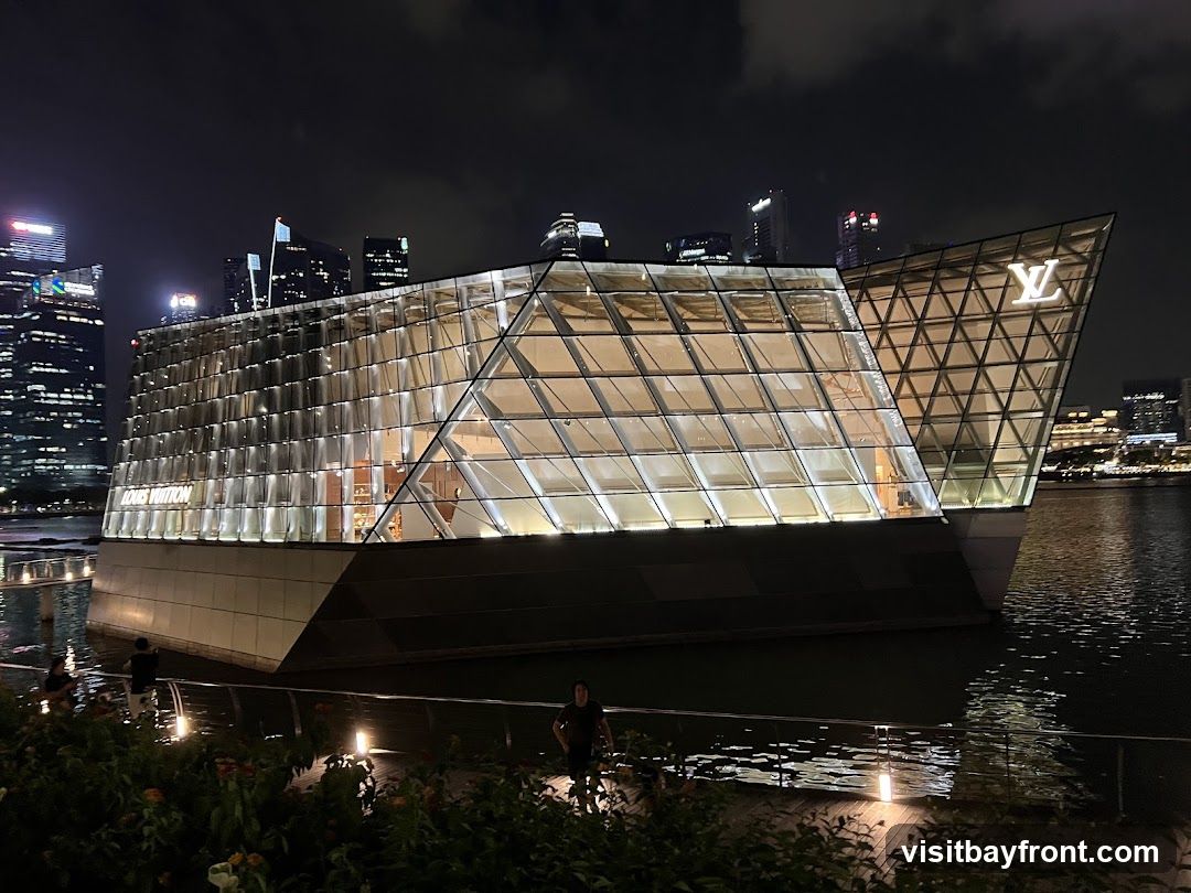 A modern, glass-encased building with a distinctive geometric design, illuminated at night. The Louis Vuitton logo is prominently displayed on one of the angled facades. The building appears to be situated on the water, with a cityscape visible in the background.