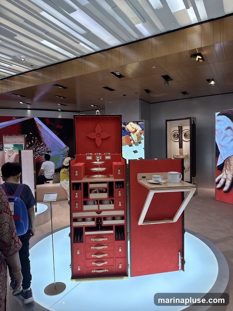 A red Louis Vuitton trunk is open, showcasing an organized interior with compartments for various items, including dishes and drawers. A small fold-out shelf displays a teacup and saucer. In the background, other displays and people are visible within a modern retail space with a distinctive ceiling design.