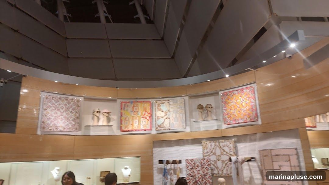 A display of luxury scarves with intricate patterns, some framed and hung on a wall, others folded and displayed on shelves. Mannequin heads are used to showcase some of the scarves. The interior features warm wood paneling and modern lighting.