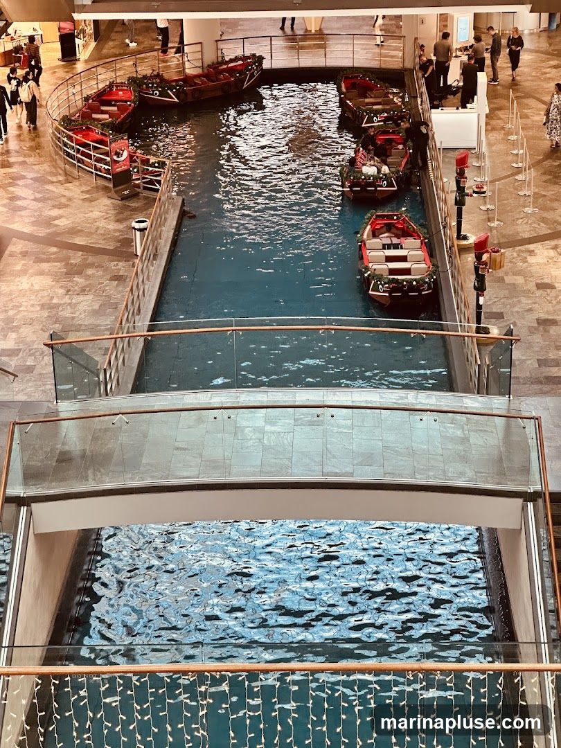 An indoor canal with several gondola-style boats decorated for the holidays. People are seen walking around the canal on the upper levels of the building. The water is a vibrant blue, and the boats are red with festive greenery.
