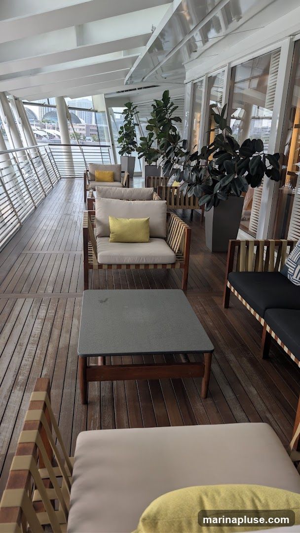 An outdoor seating area with wooden decking and modern furniture. Several chairs and sofas with light-colored cushions and yellow accent pillows are arranged around coffee tables. Large potted plants add greenery to the space. The area is covered by a sloped roof with a view of buildings and water in the background.