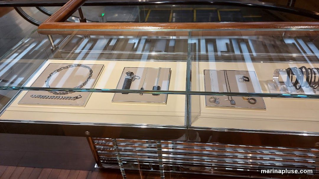 A close-up view of a well-lit jewelry display case. The case features multiple compartments showcasing various pieces of jewelry, including necklaces, bracelets, rings, and what appear to be pendants or charms. The jewelry is arranged on light-colored velvet or satin stands within glass-enclosed sections. The overall aesthetic is sleek and modern, with polished metal accents on the display case itself. In the background, reflections suggest an indoor environment, possibly a mall or department store, with escalators visible.