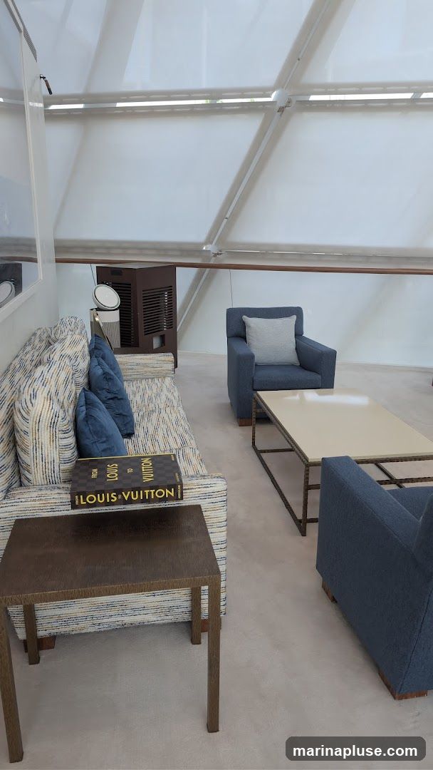 A modern living room area with blue upholstered chairs and a sofa. A coffee table sits in front of the chairs, and a side table is next to the sofa. A book with "LOUIS VUITTON" visible on its spine rests on the sofa. The room has a unique ceiling structure with white translucent panels.