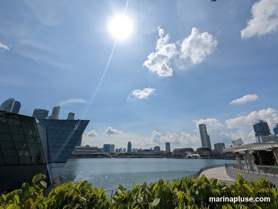 A wide shot of a modern, angular building with a Louis Vuitton logo visible on its glass facade, situated by a body of water. In the background, a city skyline with various skyscrapers and distinctive buildings rises under a bright, sunny sky with scattered clouds. Lush green foliage is in the foreground, framing the scene.